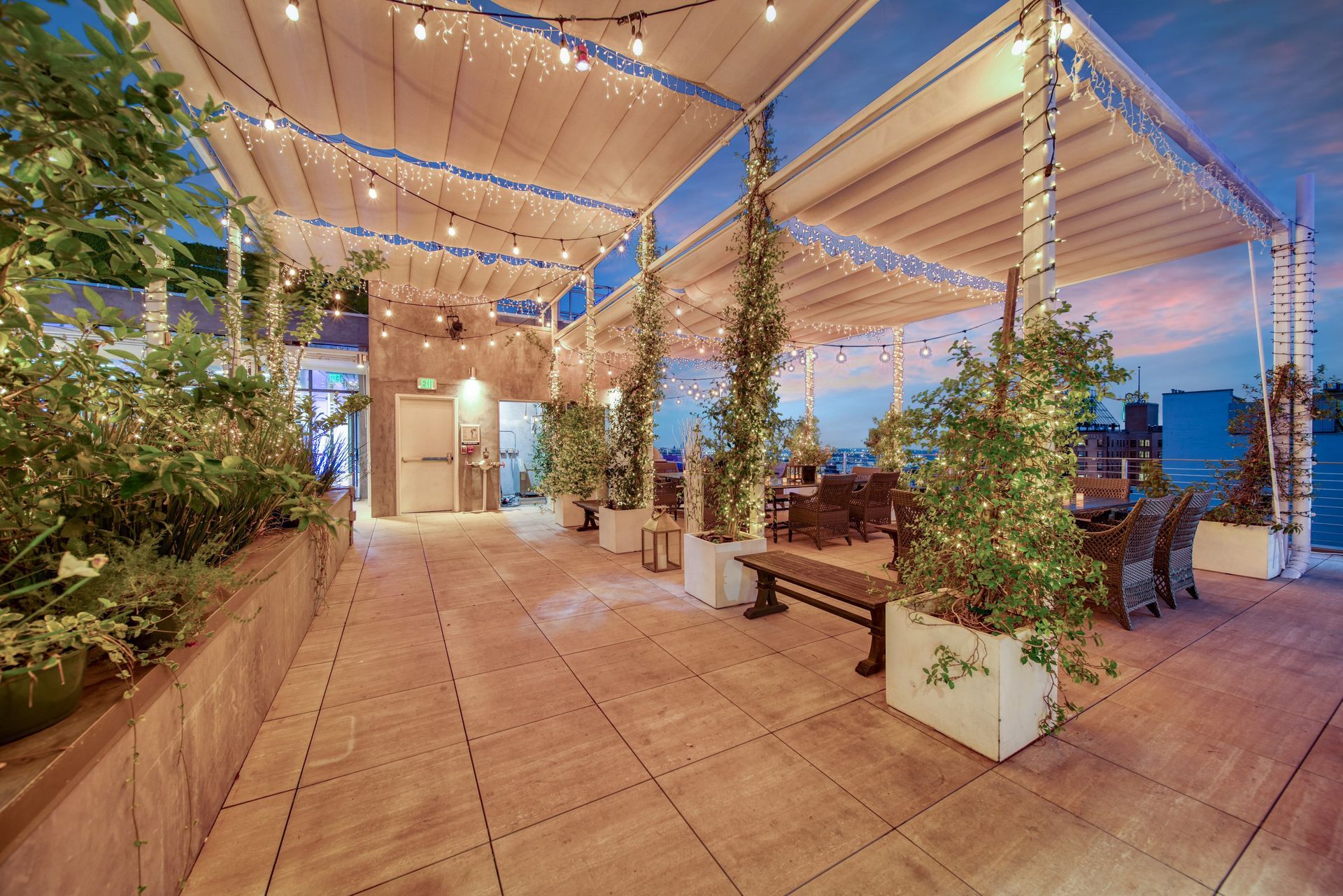 An outdoor rooftop patio at twilight featuring wood-tiled flooring, trellised plants, string lights, and bench seating.