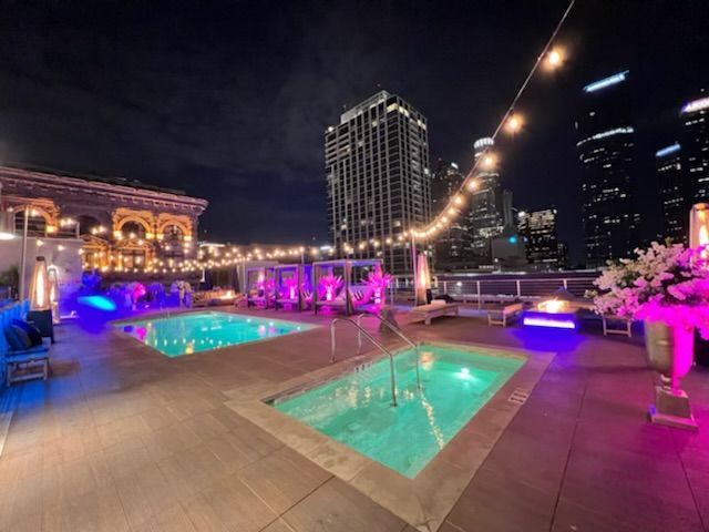 A nighttime rooftop pool deck in a city, featuring two glowing blue pools, string lights, and skyscrapers in the distance.