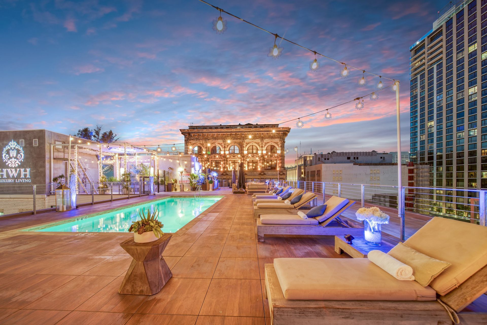 A rooftop pool with lounge chairs at sunset, featuring string lights above and a modern city skyline in the background.