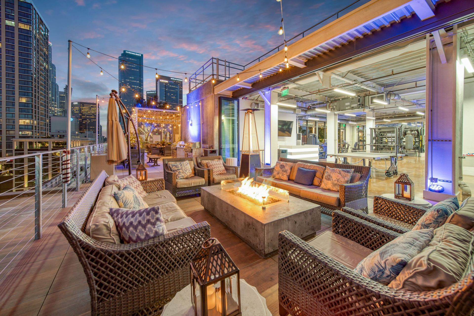 A modern rooftop patio at sunset, featuring comfortable wicker lounge furniture surrounding a stone fire pit.