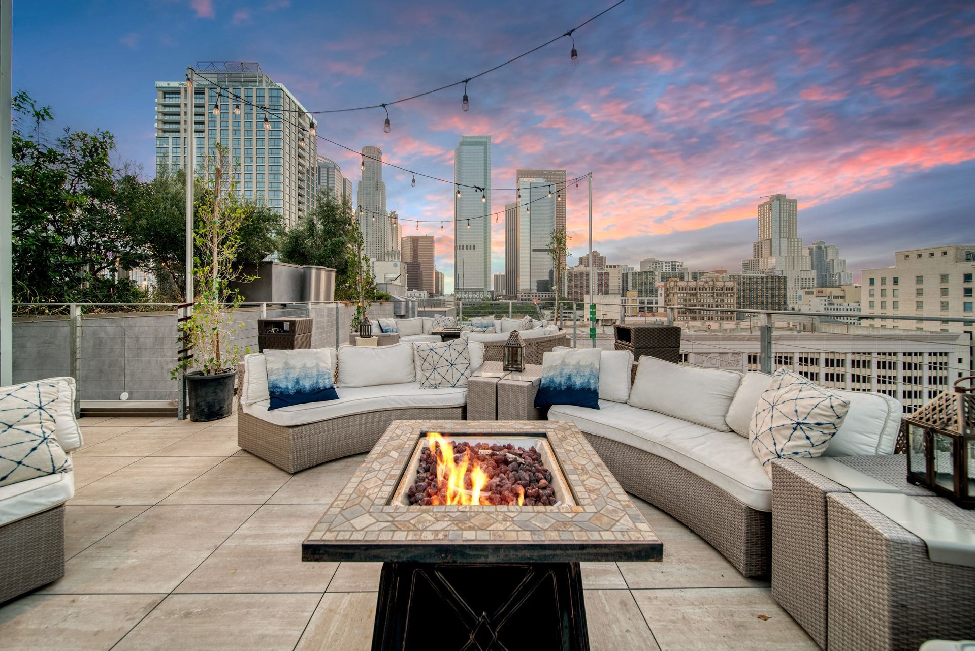 Rooftop patio with a fire pit, curved outdoor sofas, and string lights overlooking a cityscape at sunset.