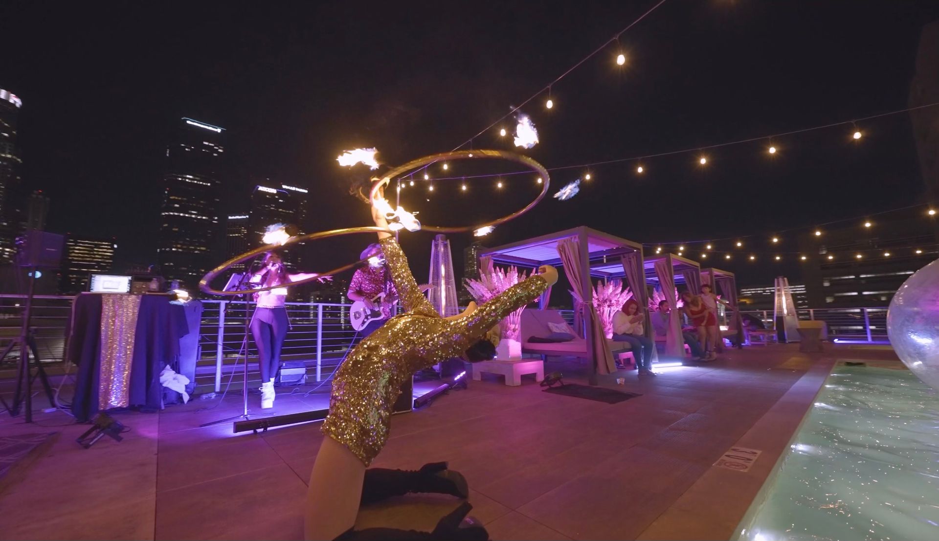 A performer in a gold sequined outfit spins a lit hula hoop on a rooftop pool deck at night, with city lights in the back.