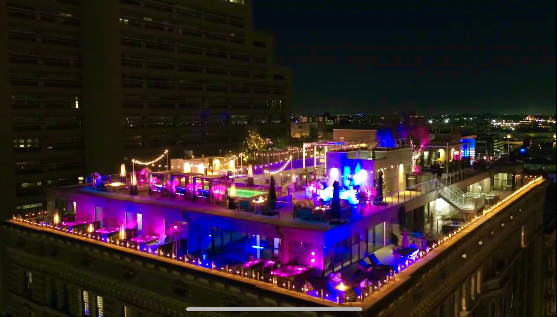 A rooftop terrace at night with bright neon purple and blue lighting, string lights, and an outdoor lounge area.