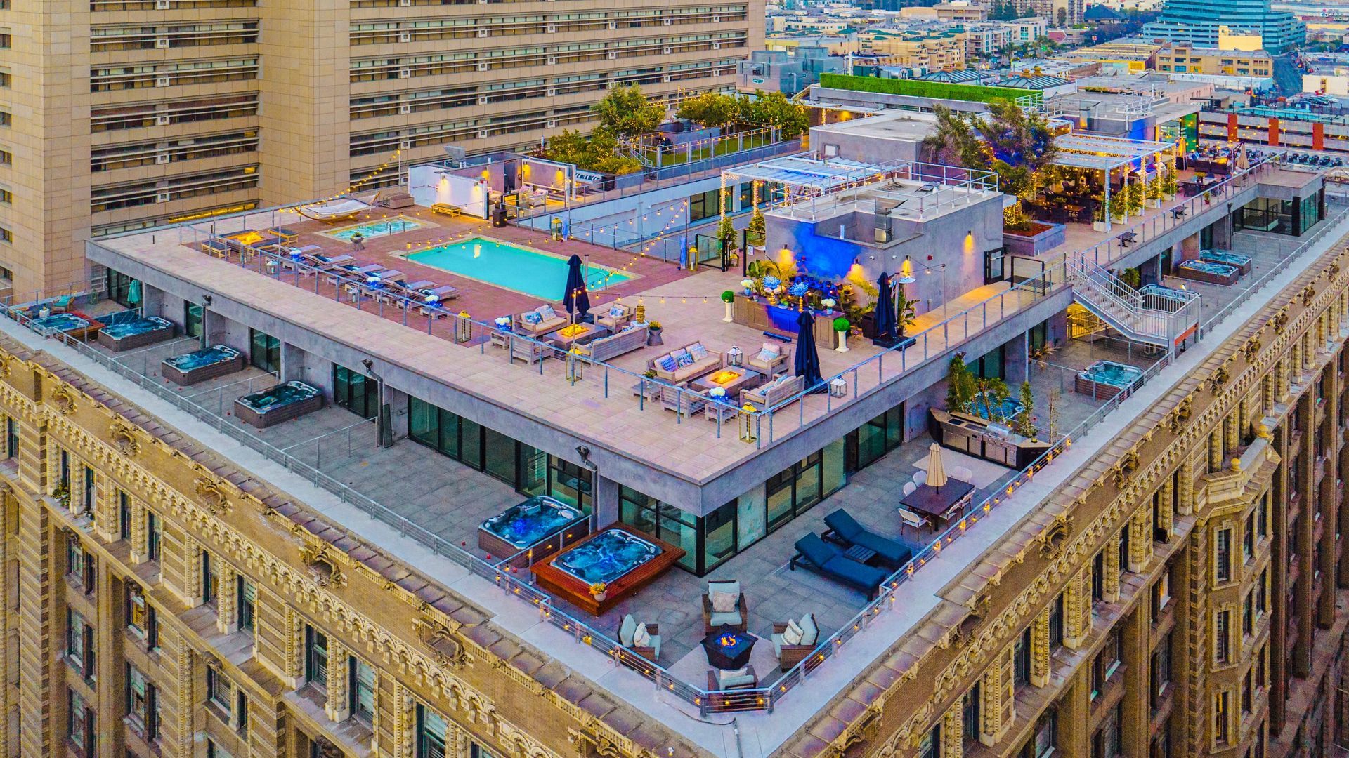 Aerial view of a rooftop hotel terrace featuring a swimming pool, lounge chairs, and hot tubs on a city building.