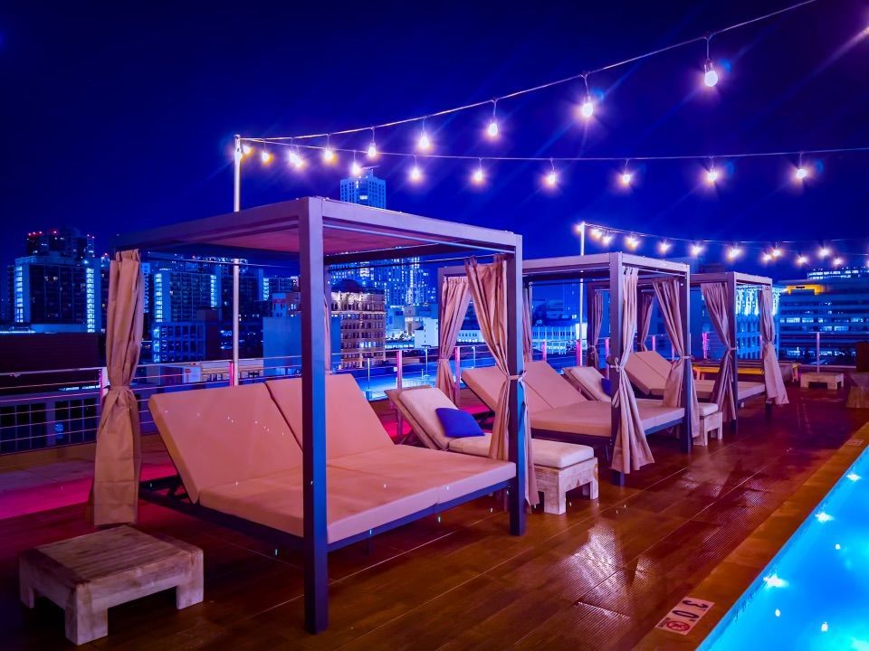 Outdoor rooftop lounge with canopy daybeds next to a lit swimming pool under string lights at night.