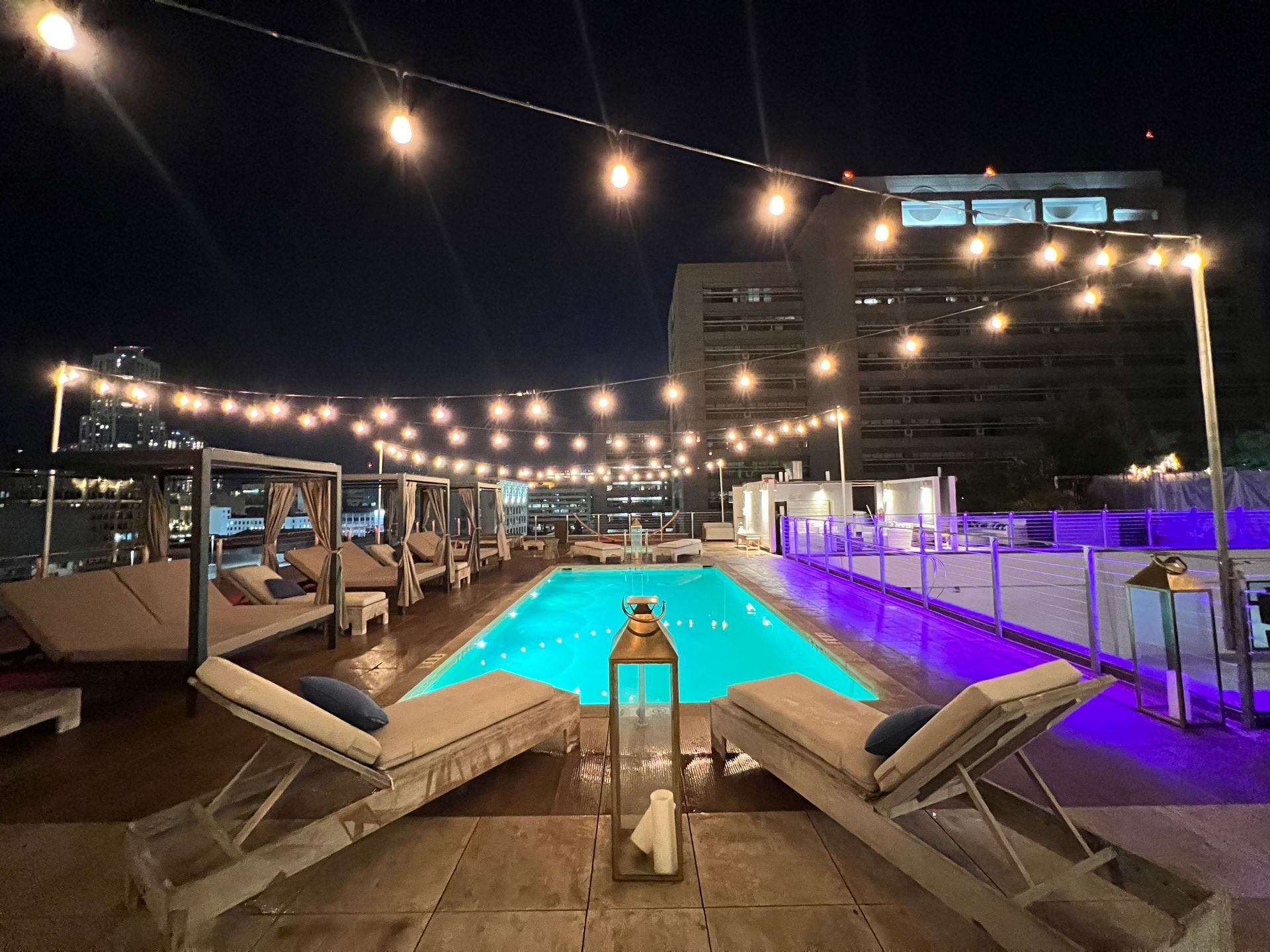 Nighttime view of a rooftop swimming pool with string lights, lounge chairs, and cabanas under a dark sky.