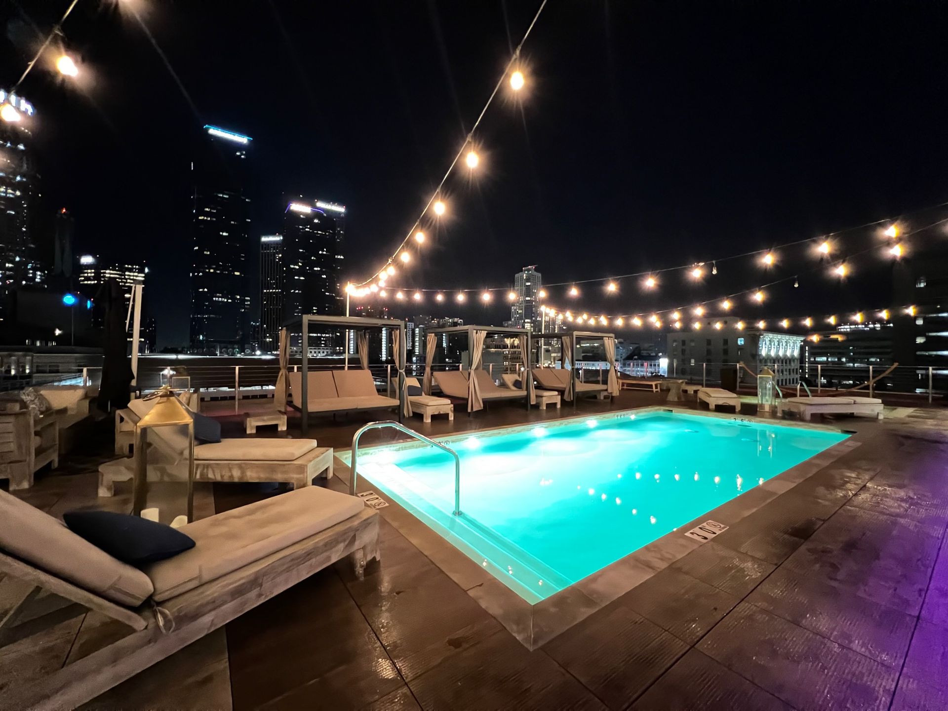 A rooftop pool at night illuminated by string lights, surrounded by lounge chairs and cabanas with city skyline views.