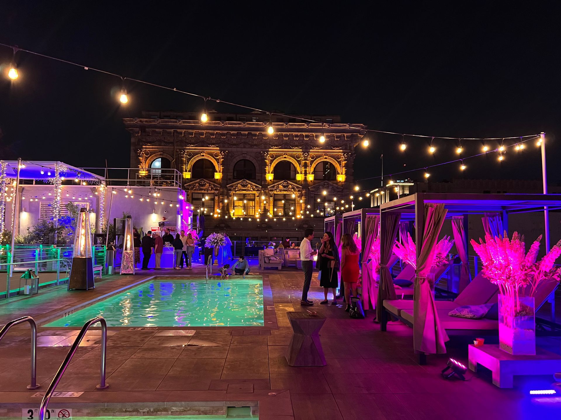 A brightly lit outdoor pool deck at night, featuring a swimming pool, lounge cabanas, and glowing string lights.