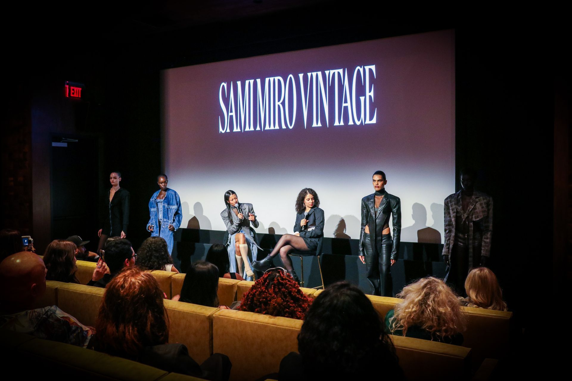 A fashion presentation on stage at a theater with models standing beside two seated people in front of a brand logo.