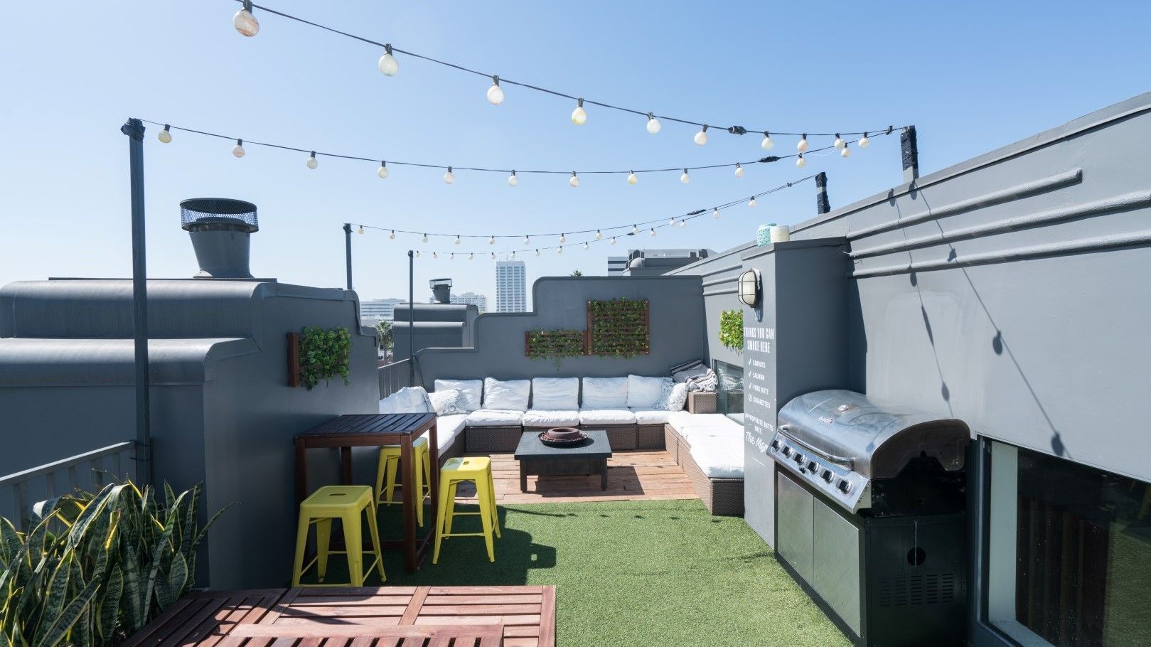 A rooftop deck with a couch , table , stools and a grill.