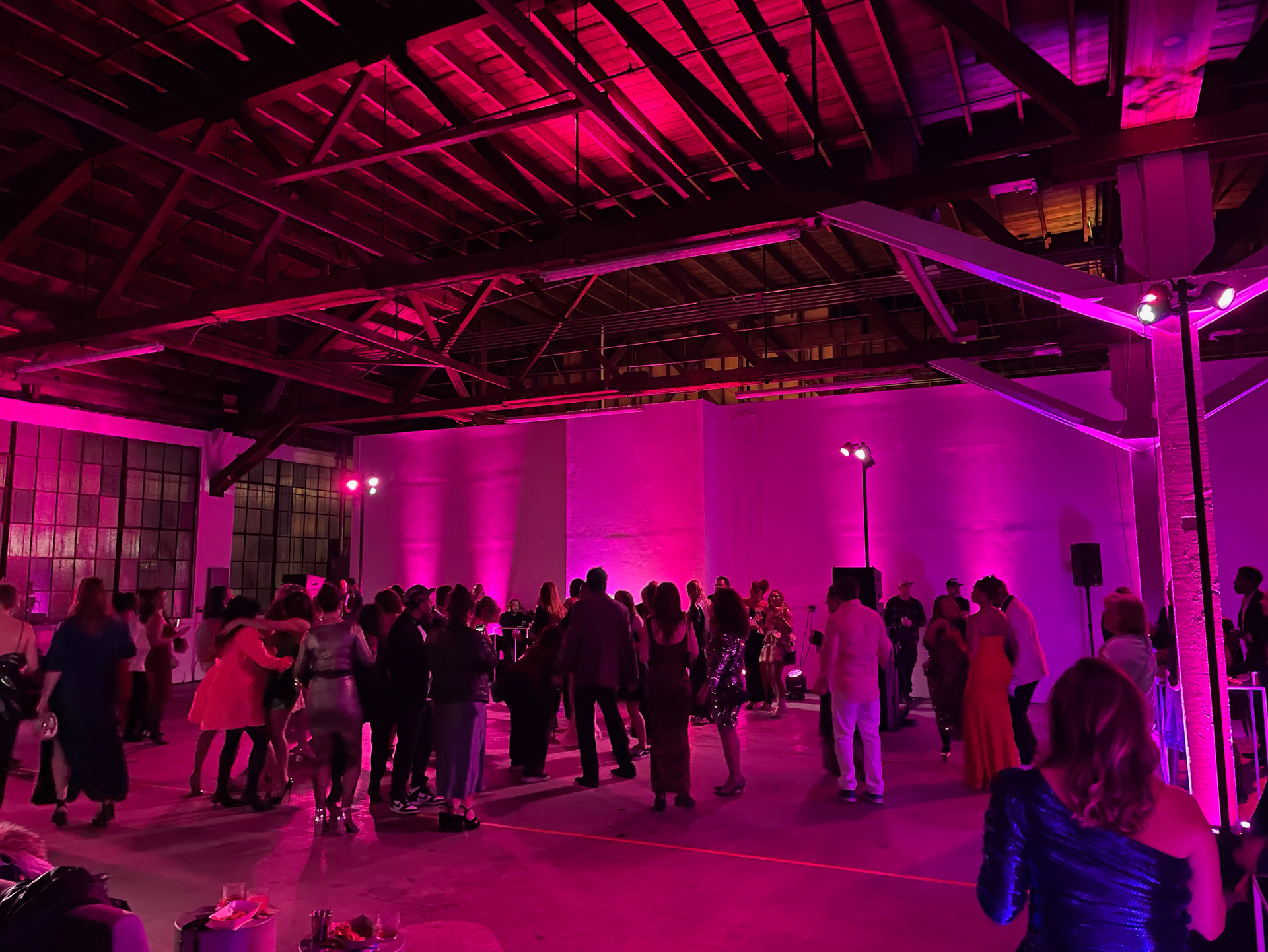 A group of people dances in an industrial-style hall illuminated by vibrant pink lighting.