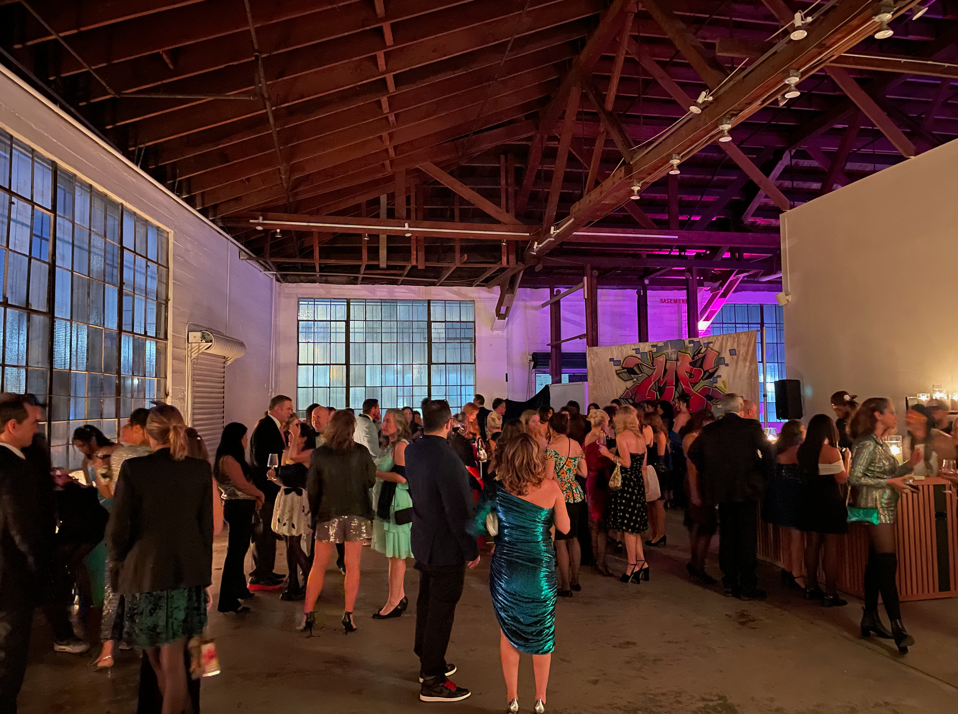 A large, industrial venue with exposed wooden beams, filled with people in cocktail attire attending an indoor event.