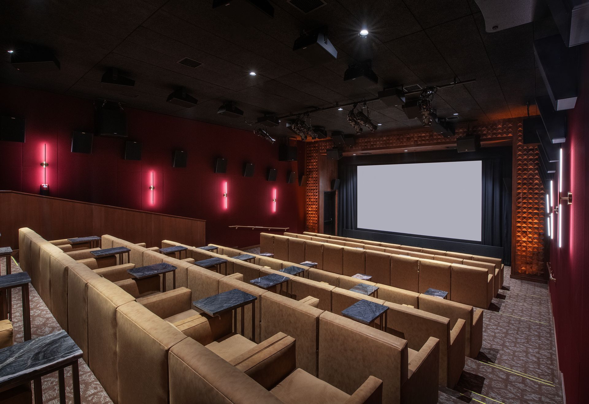 A dim, modern movie theater with rows of tan leather armchairs, marble side tables, and vertical red accent lighting.