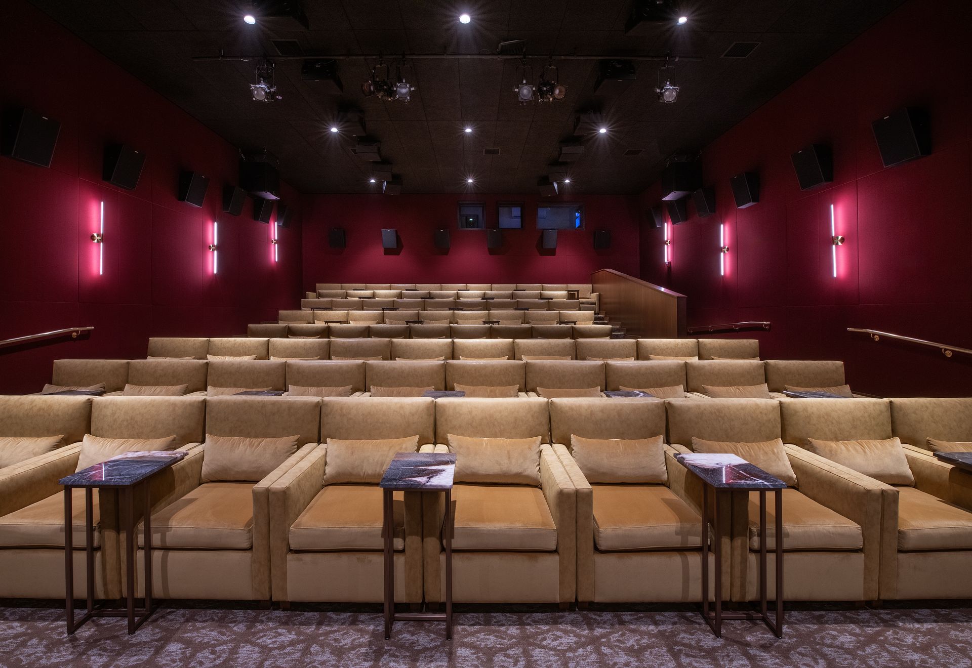 A movie theater with rows of tan cushioned armchairs, small side tables, and red walls with accent lighting.