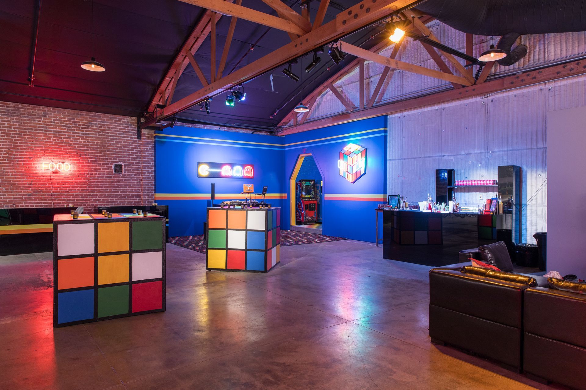 A brightly lit game room featuring Rubik's Cube-themed tables, neon signs, and black leather sofas on a concrete floor.