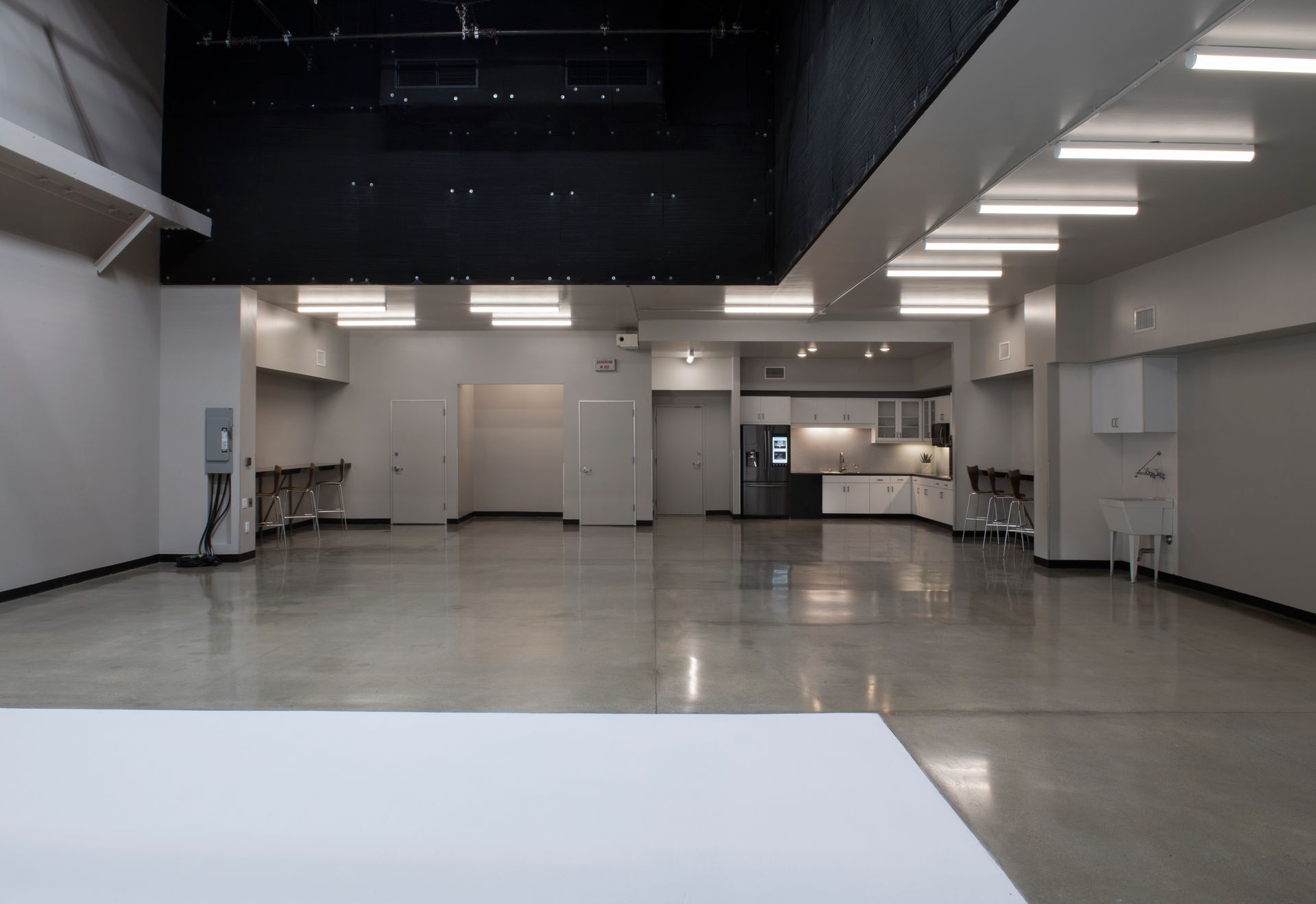 A wide, modern room with light gray walls, polished concrete floors, recessed lighting, and an open kitchen area.