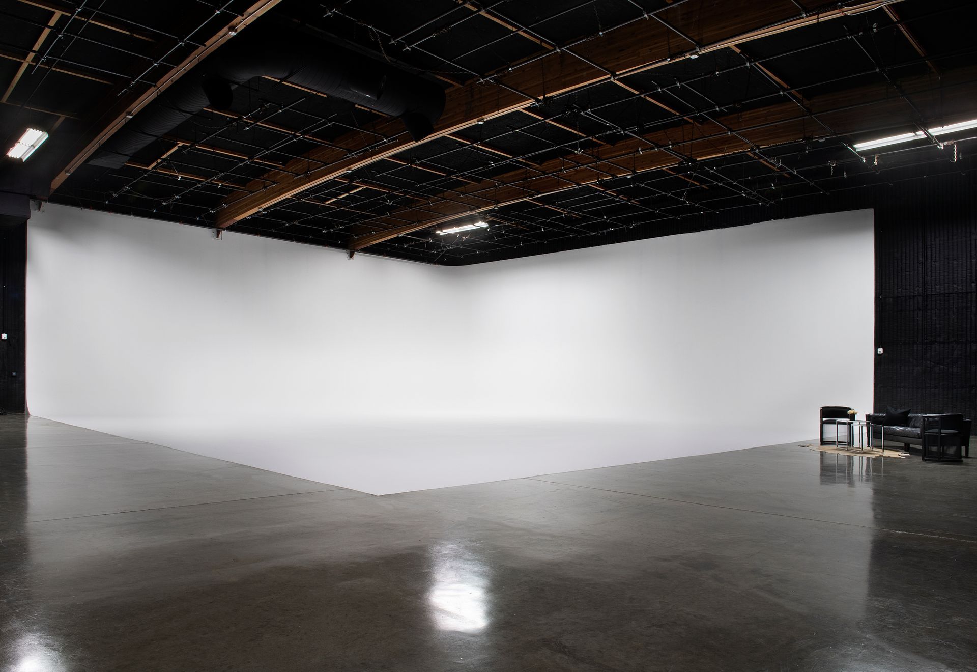 A large, empty studio with a white cyclorama wall, high black ceilings with beams, and a small black sofa in the corner.