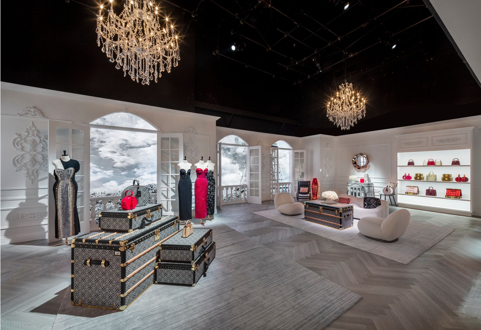 A luxury boutique interior featuring designer trunks, mannequins in evening dresses, two chandeliers, and a bag display.