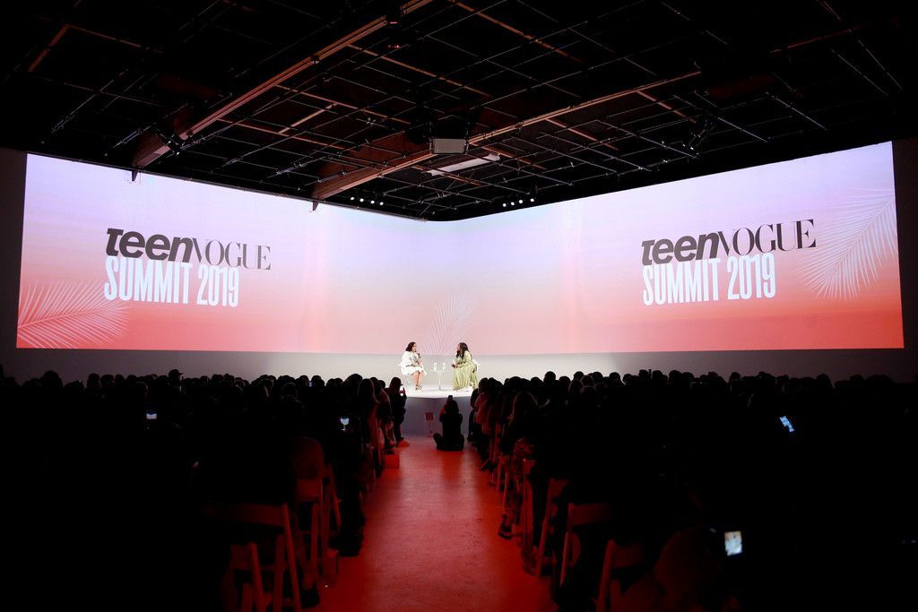 Two people on a stage at the Teen Vogue Summit 2019, set against a large dual-screen backdrop in a dark, crowded room.
