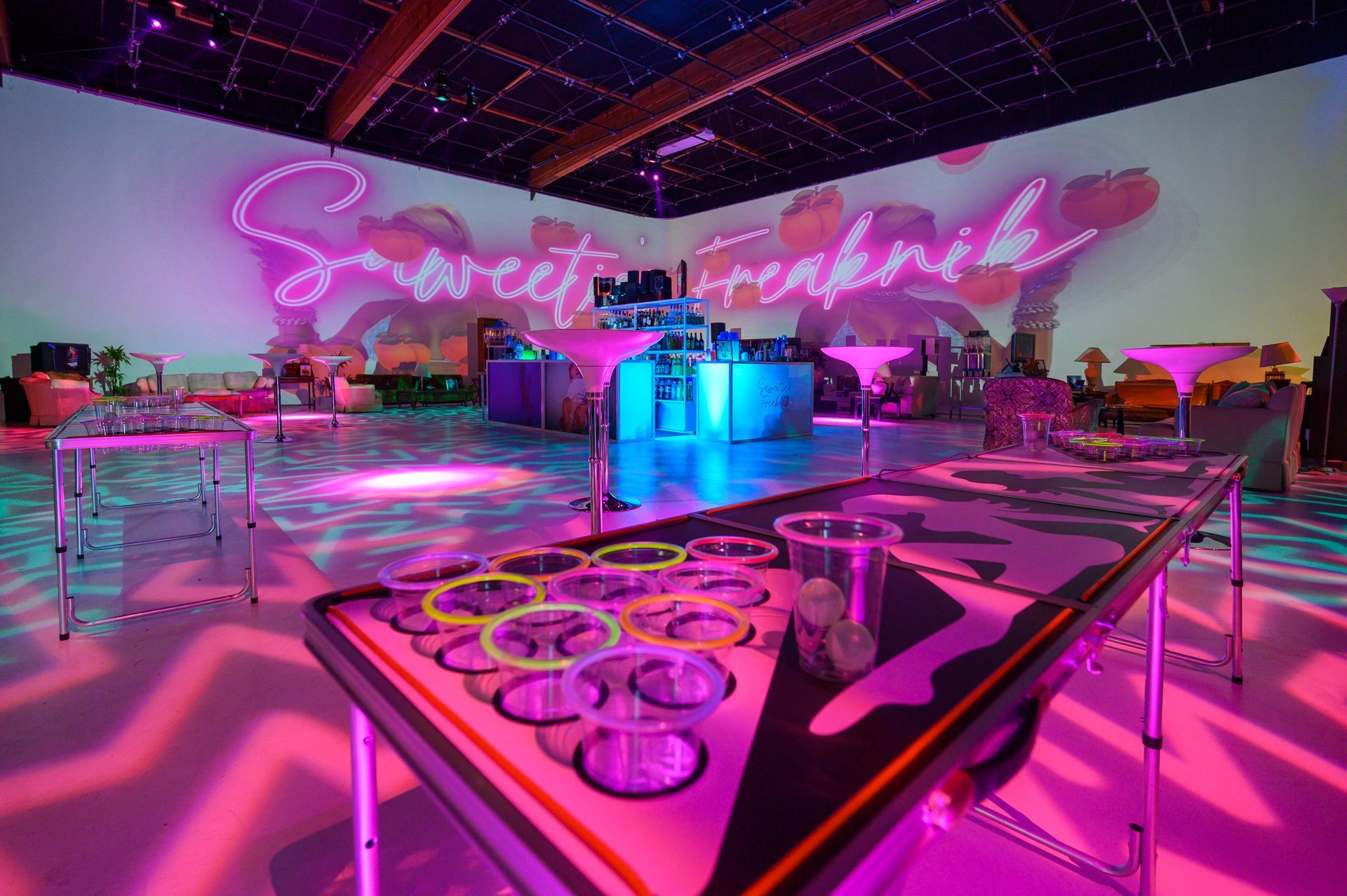 A room lit in neon pink with a beer pong table in the foreground and projection walls featuring the text 
