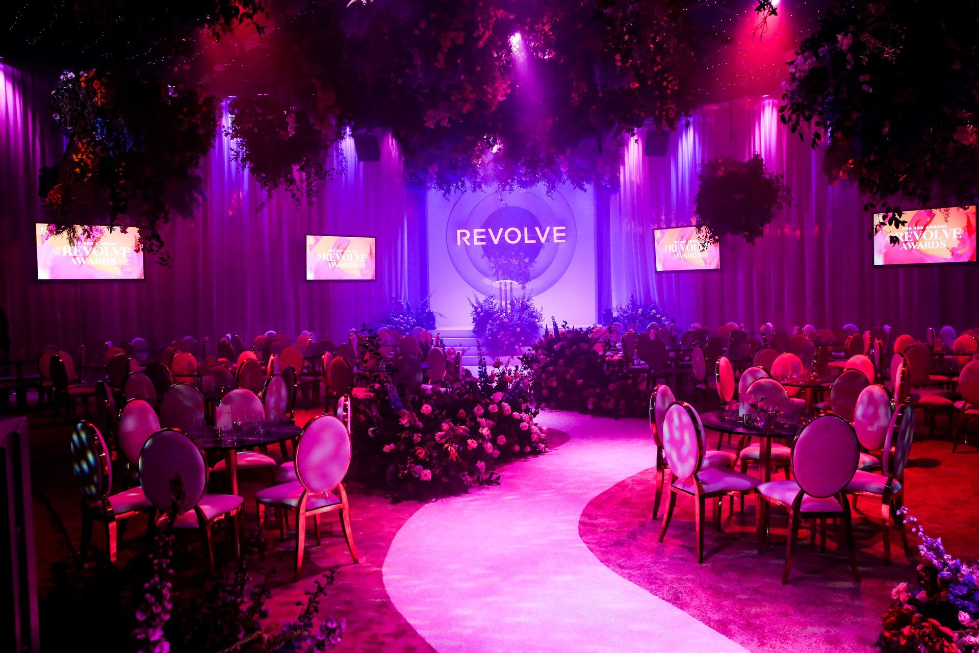 A dimly lit event space featuring a winding white path, purple lighting, round tables, and a Revolve brand backdrop.