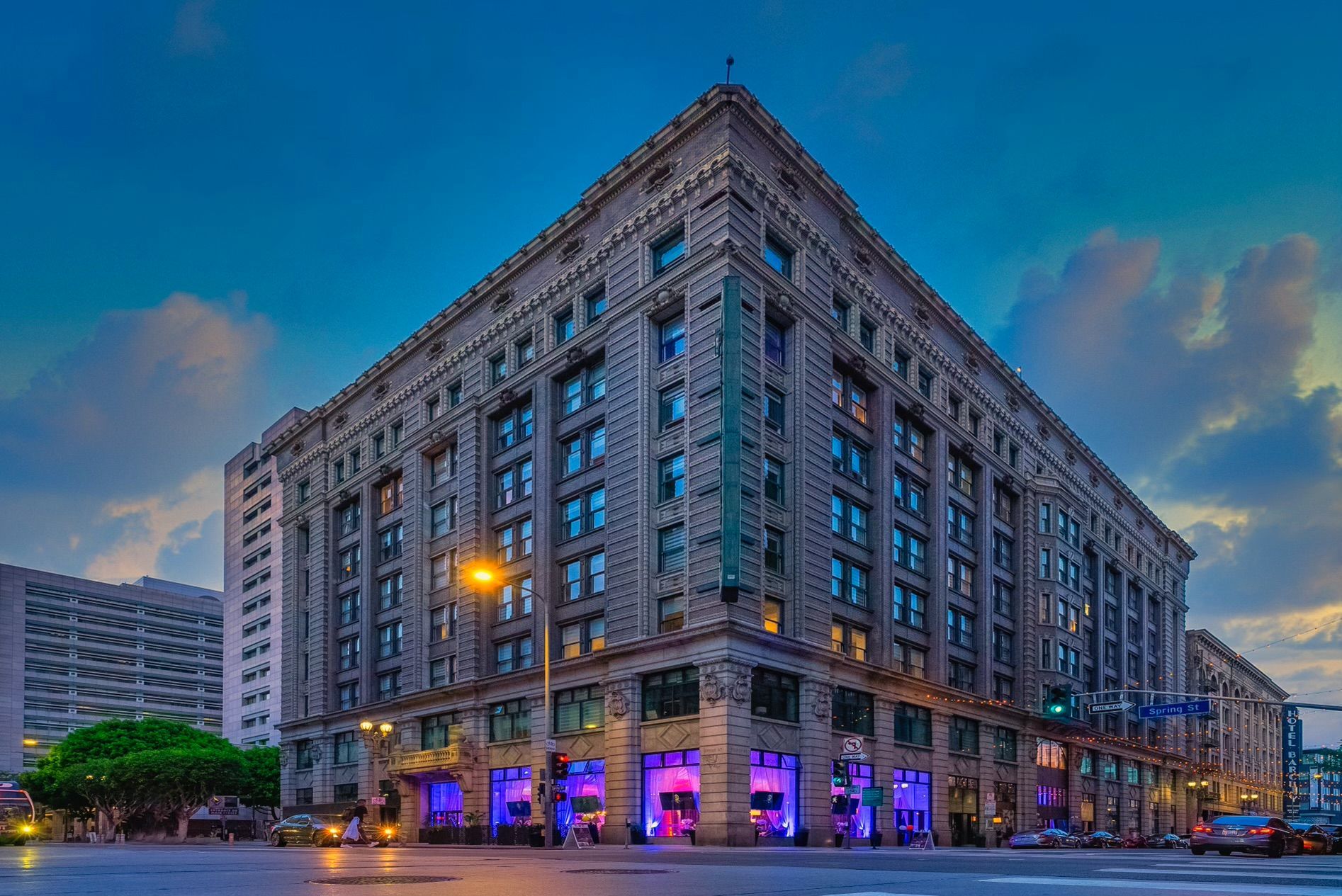 A grand, ornate stone building at a corner intersection under a dusky blue sky, featuring illuminated ground-floor shops.