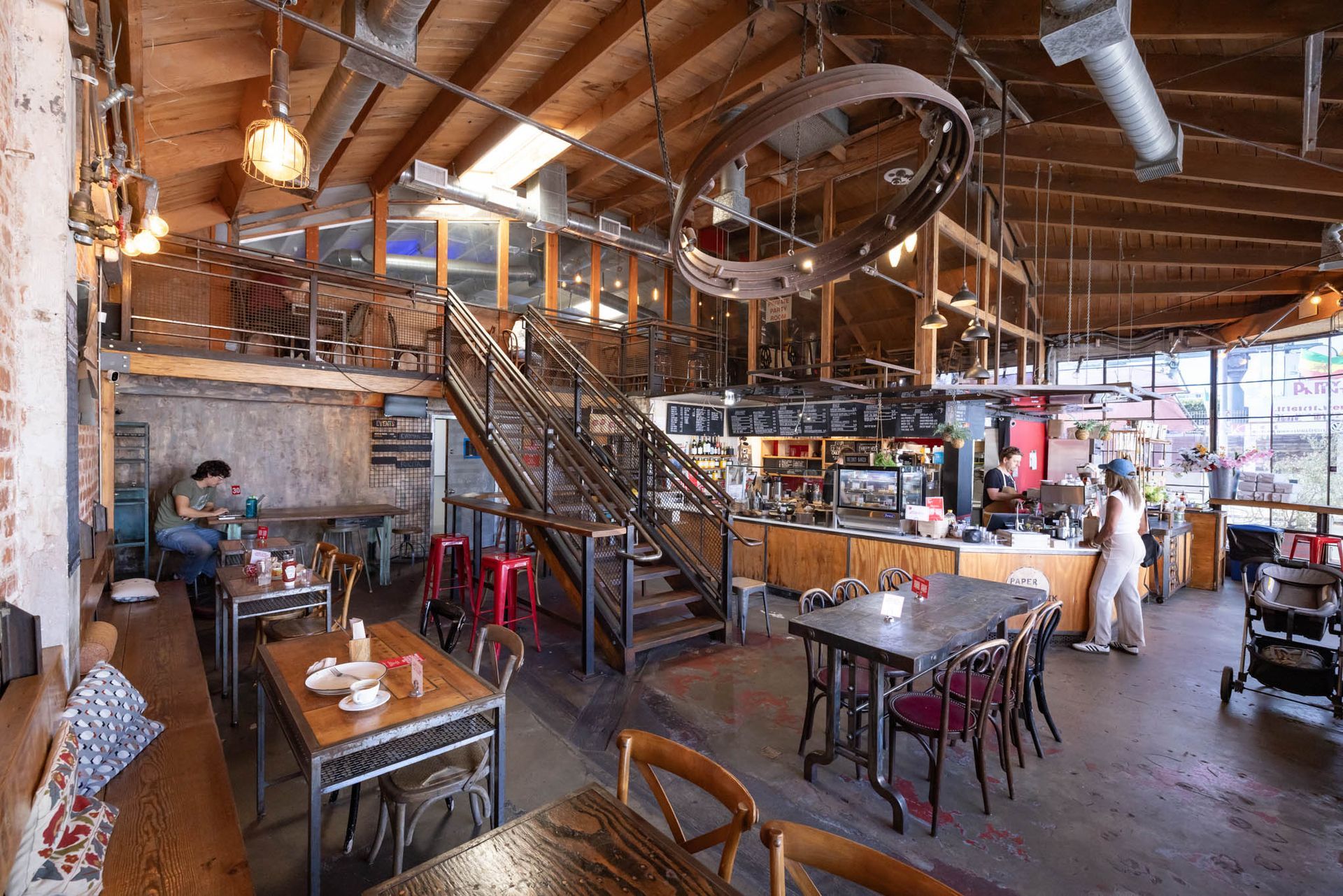 A rustic, two-story restaurant interior with industrial wooden beams, a central metal staircase, and cafe-style seating.