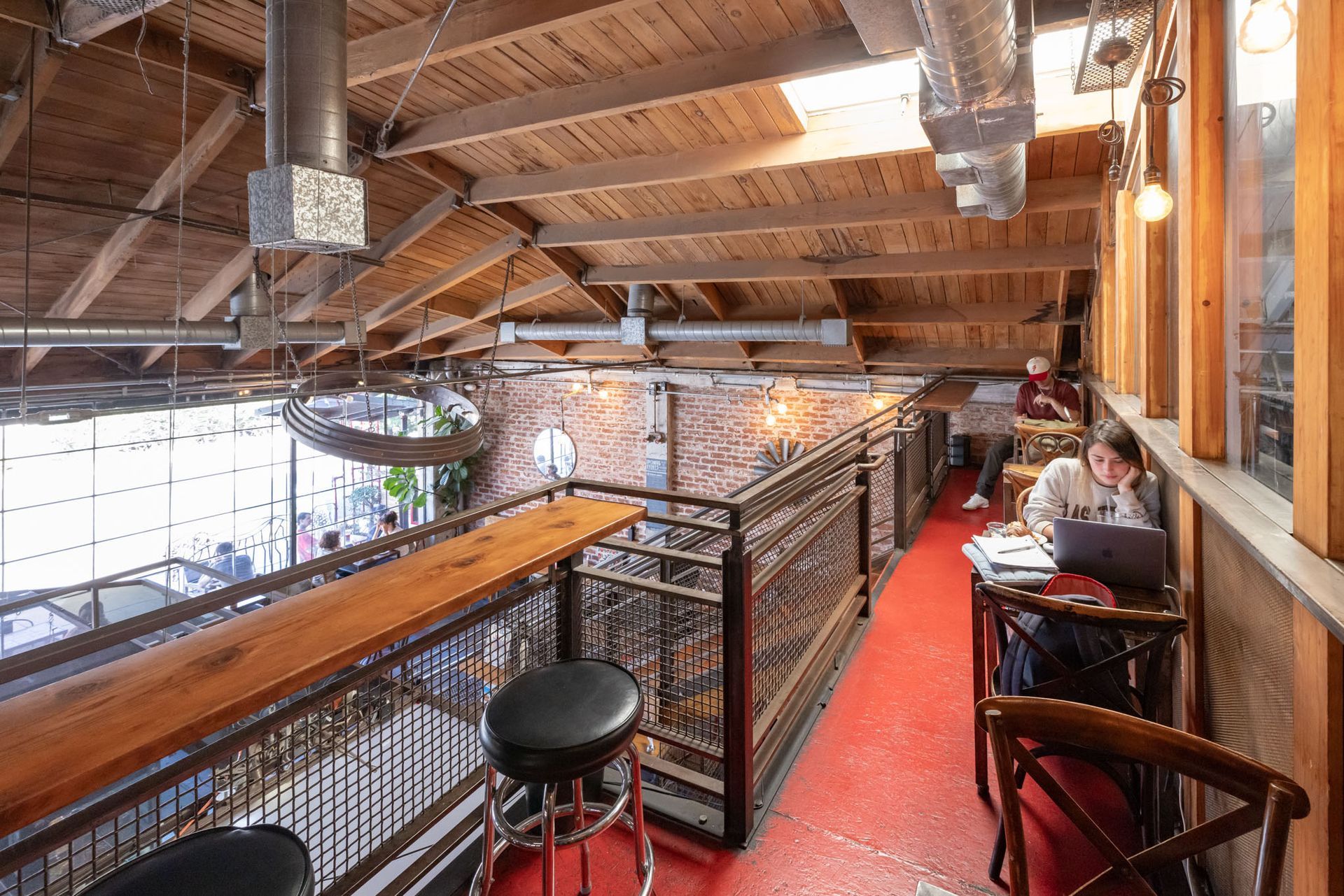 A balcony loft in an industrial-style café with a red floor, wooden ceiling, brick walls, and seating for patrons.