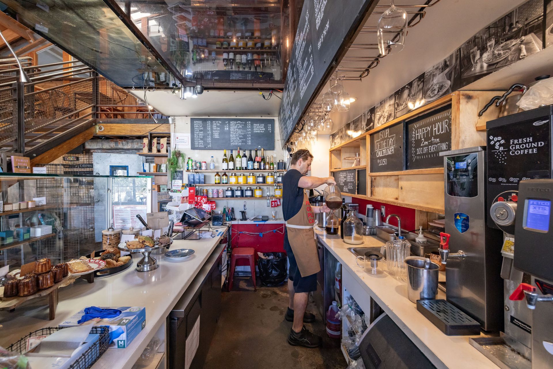 A person wearing a brown apron works behind the counter of a brightly lit, rustic-style coffee shop with menu boards.