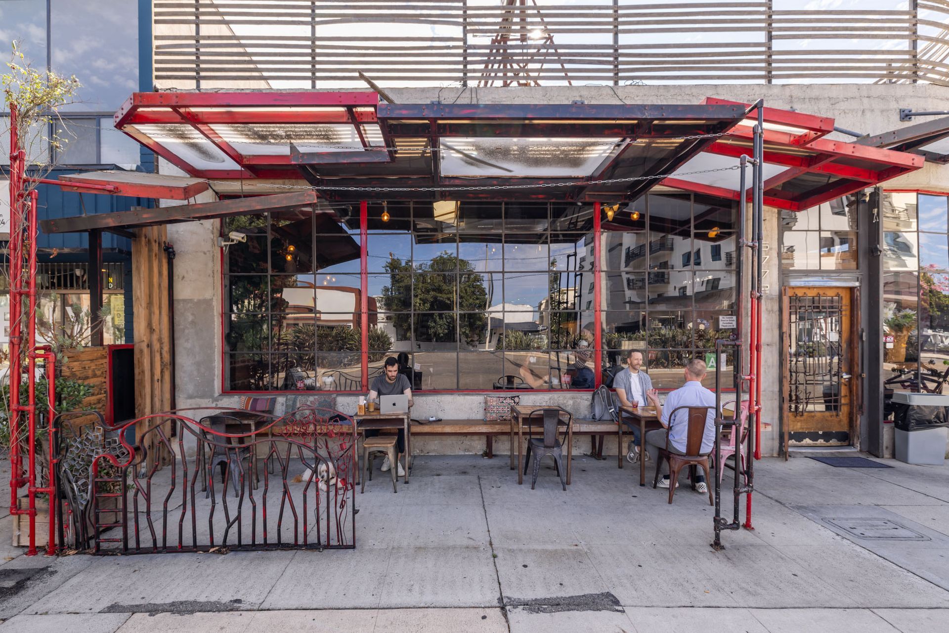 A cafe patio with red metal framing, outdoor seating, and a large windowed storefront on a paved sidewalk.
