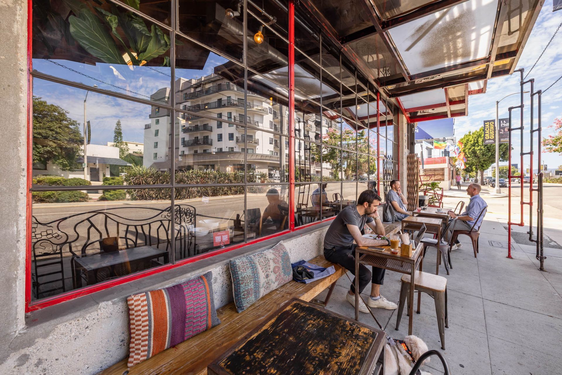 People sit at tables on an outdoor patio along a sidewalk, seen through large windows reflecting a nearby building.