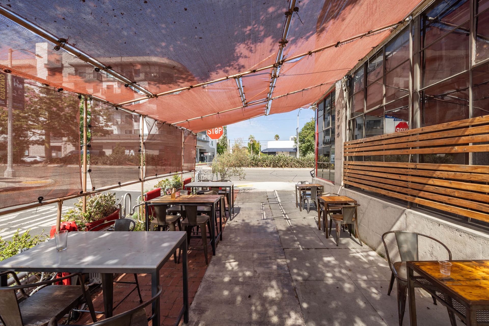 Outdoor patio seating area with metal tables and chairs shaded by a large, reddish-orange fabric canopy.
