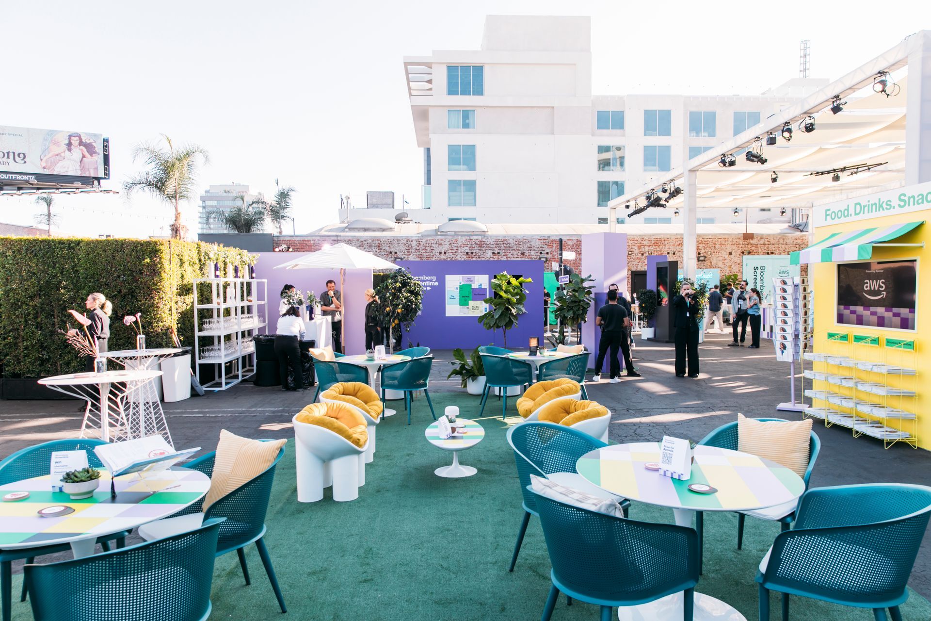 An outdoor rooftop event space with circular tables, teal chairs, and a yellow snack kiosk under a bright, sunny sky.