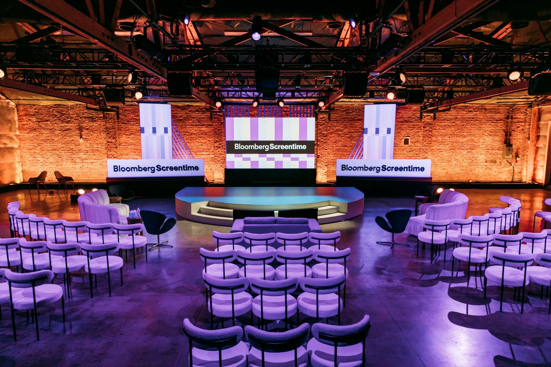 An empty indoor conference hall with rows of chairs facing a circular stage and a large screen labeled Bloomberg.