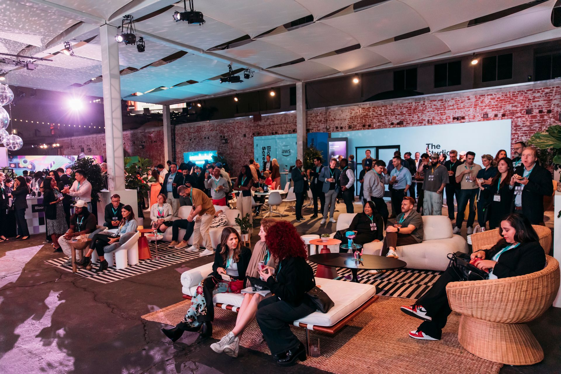 Attendees mingle and sit on modern furniture at an indoor, industrial-style social gathering with a brick wall backdrop.