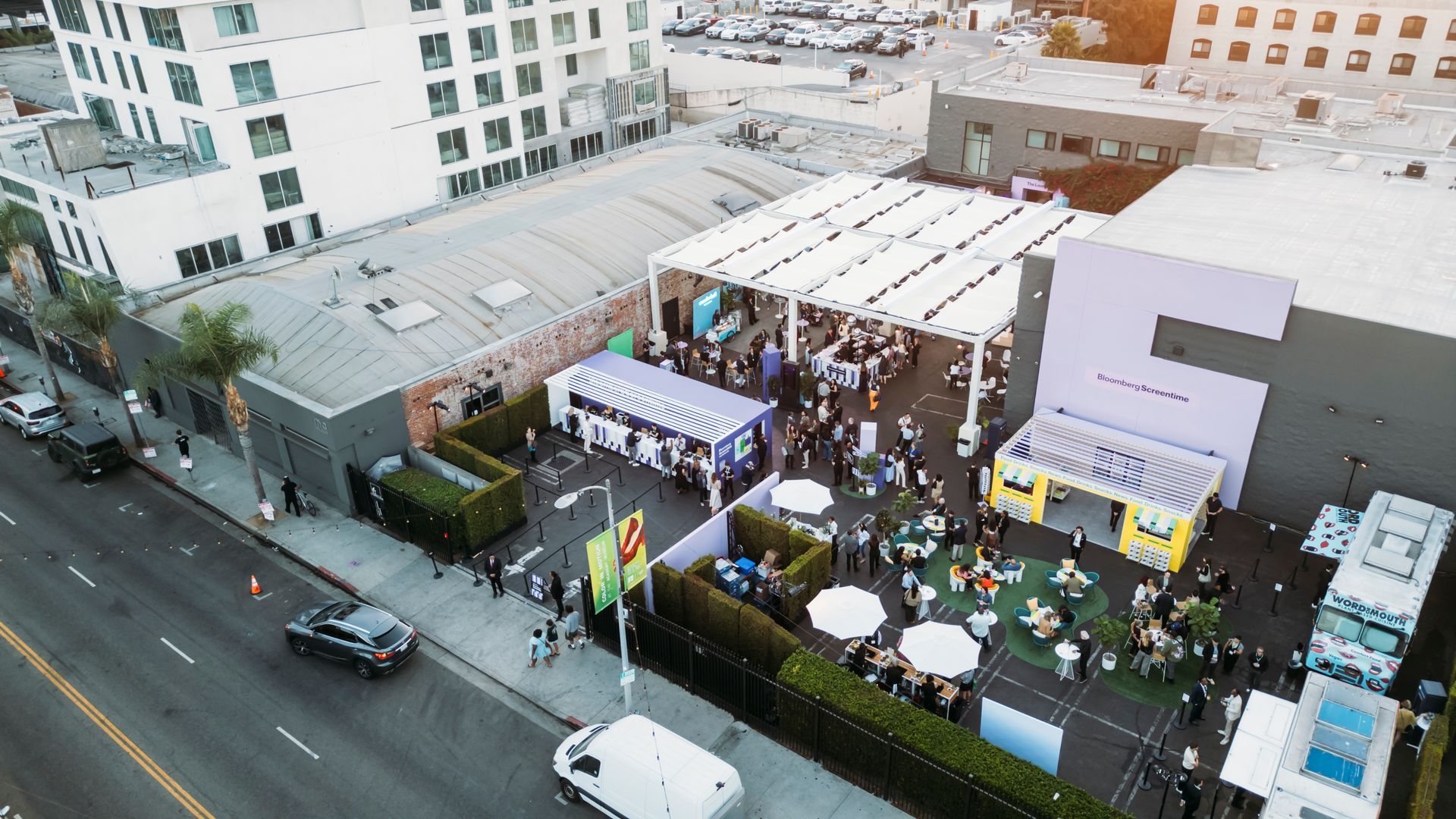 An aerial view of an outdoor event with white tent canopies, a crowd of people, and food trucks on a paved urban lot.