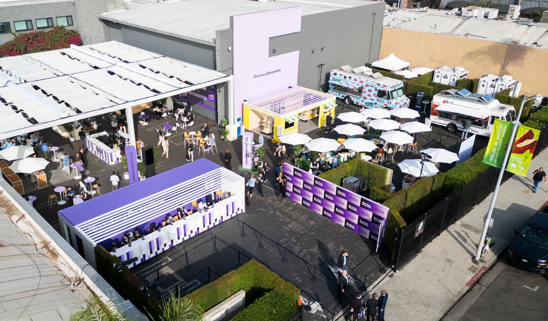 Aerial view of an outdoor event with white tents, food trucks, purple branded walls, and seating areas on a paved lot.