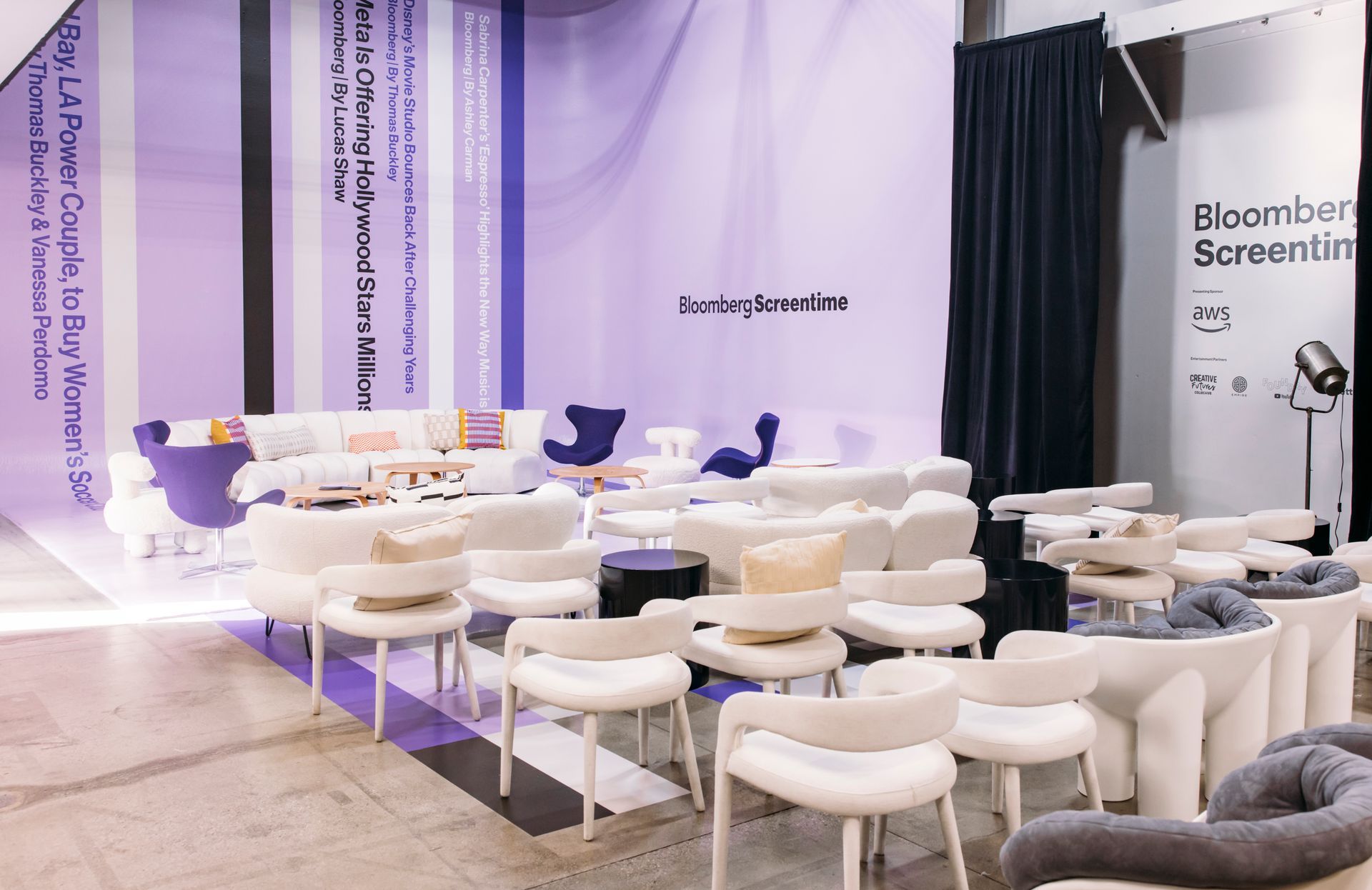 An event stage with white modern chairs and sofas arranged in a seating area against a lavender purple backdrop.