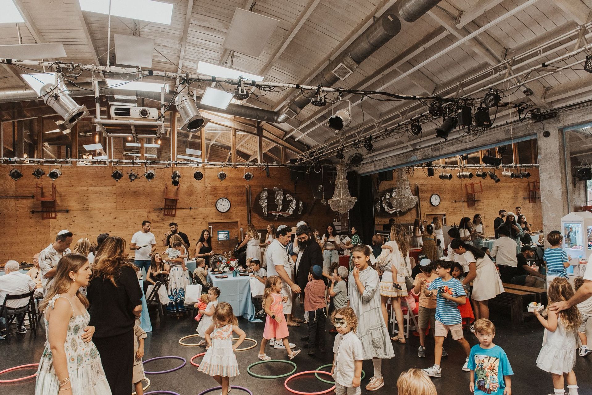 Children and adults in a rustic, industrial event space with hula hoops on the floor and high-ceiling lighting rigs.