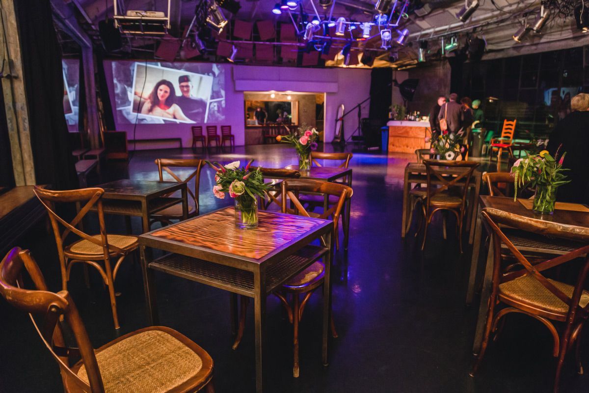 An event space set up with wooden tables and chairs, a projection screen showing a photo, and purple stage lighting.