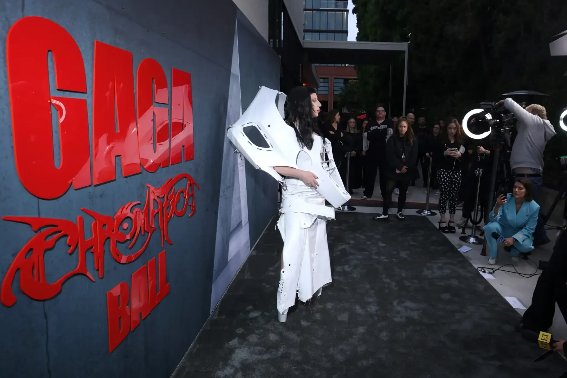 A person in a white avant-garde outfit stands on a dark carpet before a red 