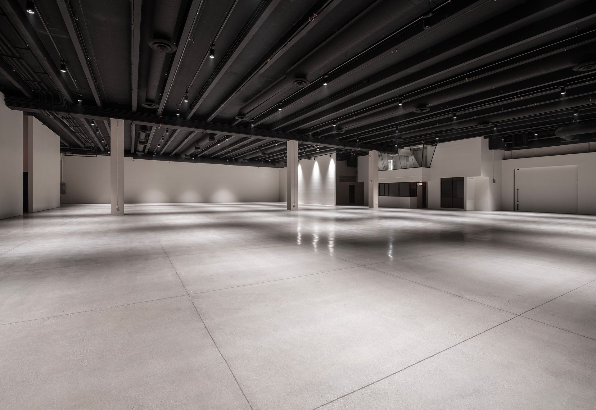 A large, empty minimalist event space with polished concrete floors, white walls, and a black industrial ceiling.