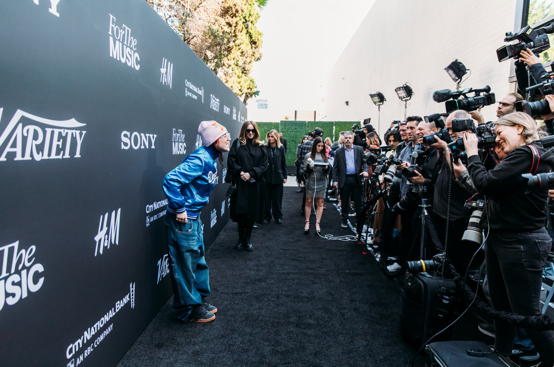 A person in a blue jacket and beanie stands on a carpeted path before a wall of photographers at a Variety media event.