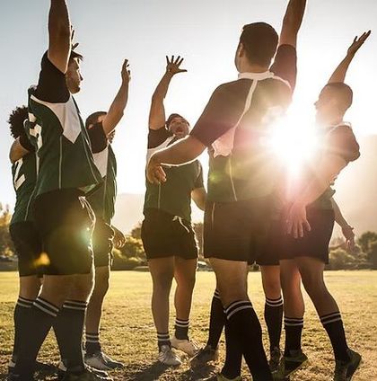 Rugby players huddle with raised hands on a sunlit field, backlit by the setting sun