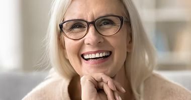Smiling woman with new full dentures her chin on her hands in a bright indoor setting