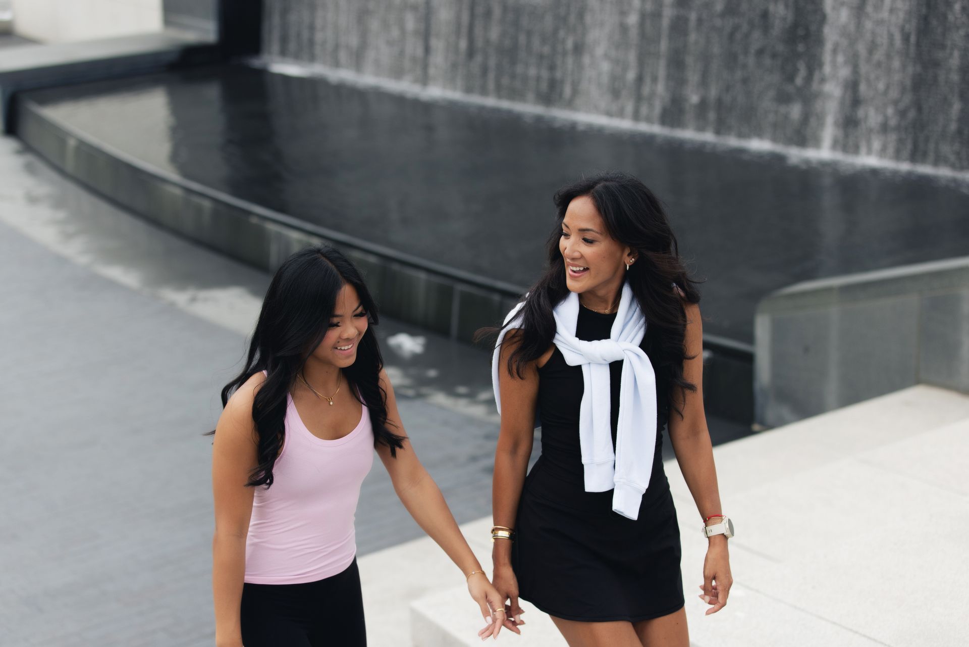 Two women smiling, holding hands, near a modern water feature. One in black dress, the other in pink.