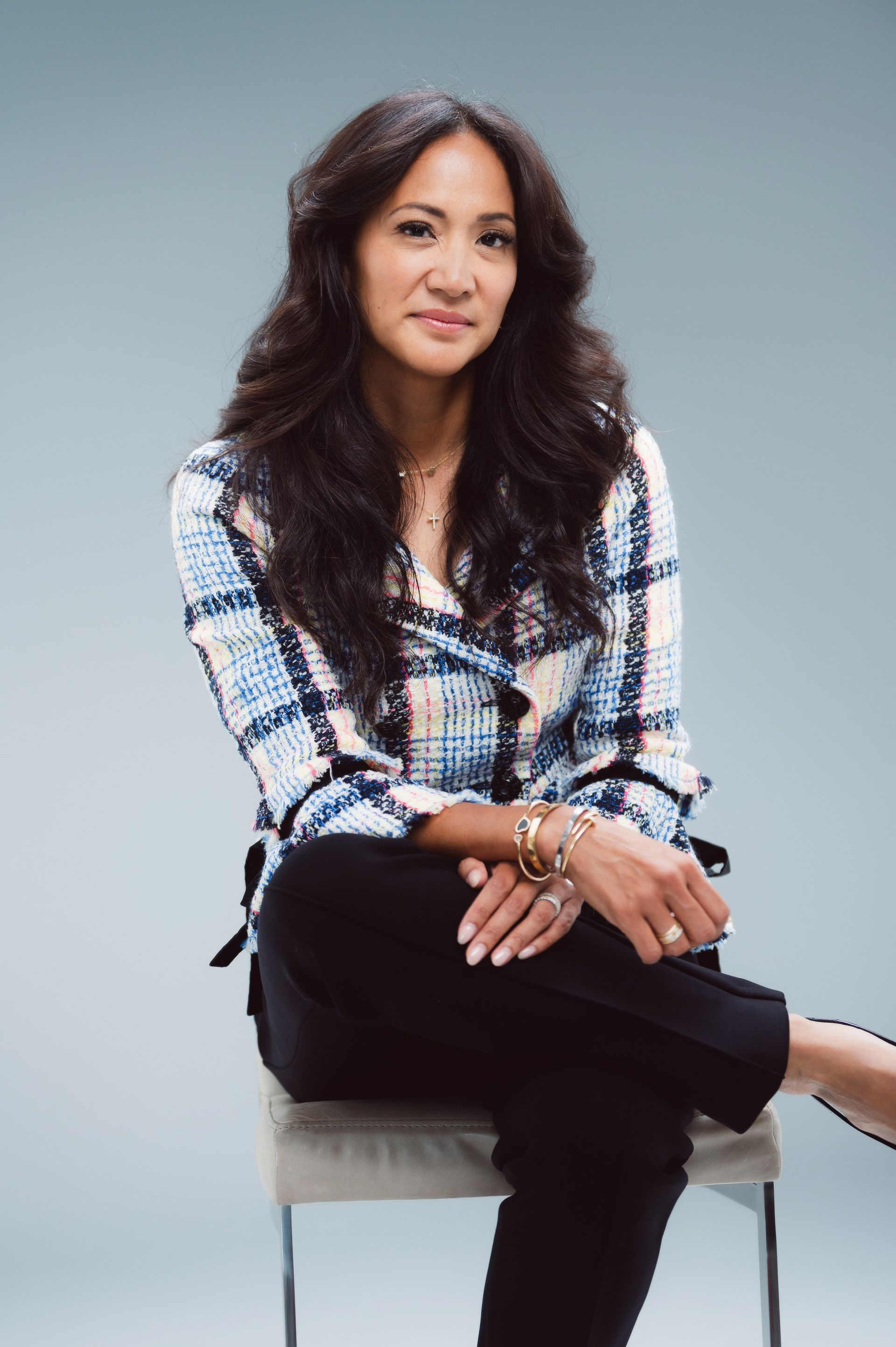 Woman seated on a stool, wearing a plaid blazer and black pants, looking at the camera.