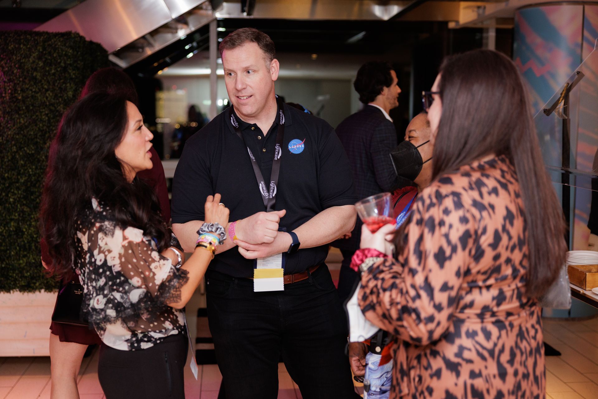 Three people at a social gathering, conversing. The man in a dark polo shirt is flanked by two women, one in a patterned top, the other in a brown coat.