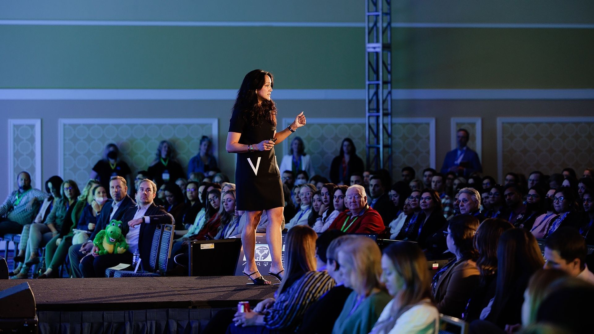 Woman in black dress giving a presentation on stage to a large audience in a brightly lit room.