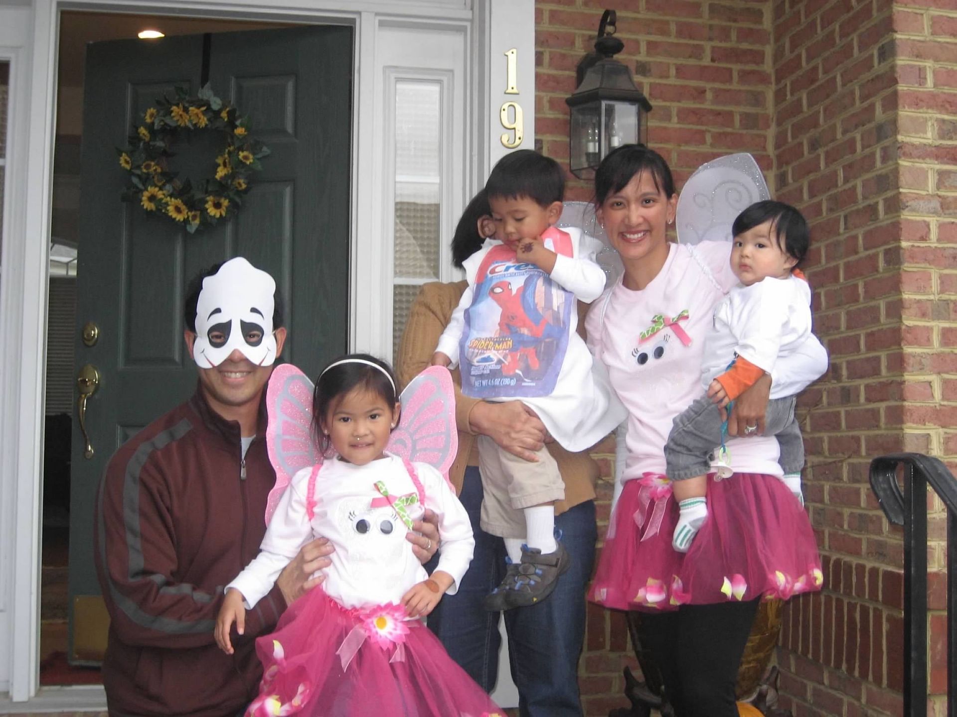 Family in Halloween costumes: dad with mask, girl fairy, mom with wings, and two toddlers.